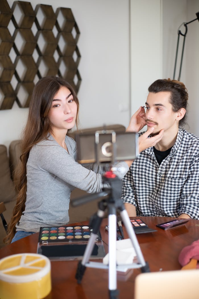 creative Asian woman applies makeup to a Caucasian man for a vlogging session, filmed using a tripod.